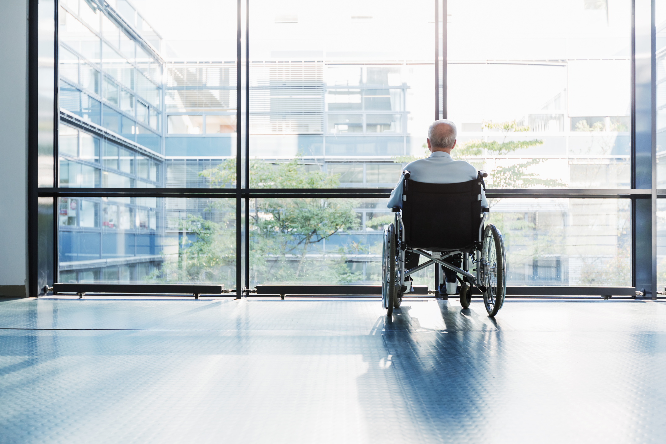 Senior Man in Wheelchair looking out of a window in a hospital corridor.